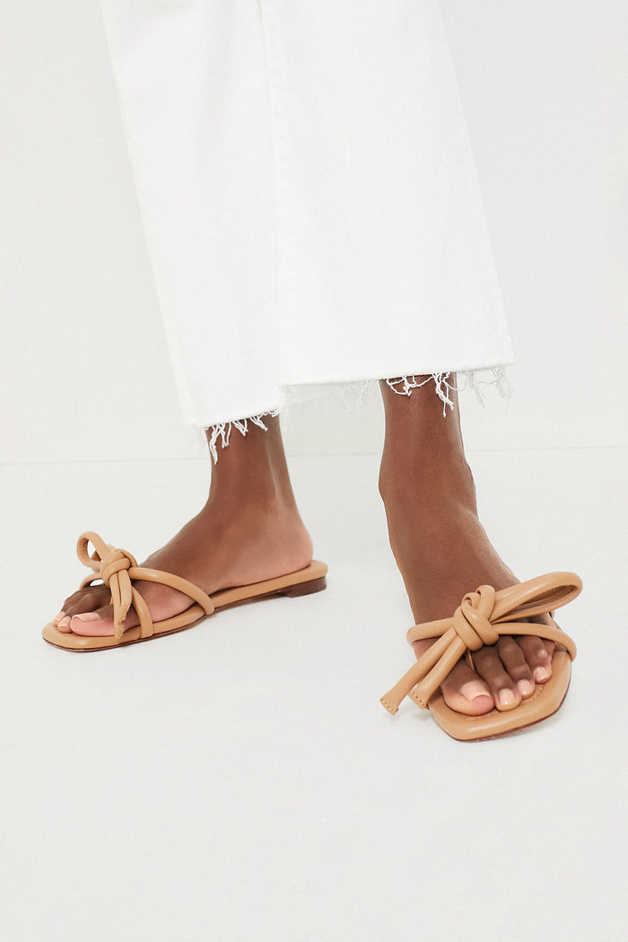 Dune Nappa Hadley Leather Bow Flat Sandal: Classic, effortless nude-tan slides with knotted straps, showcased with cropped white denim, embodying a fun, high-end style.