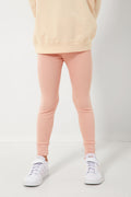 A person stands confidently in a beige Les Gamins sweatshirt, light pink Les Gamins organic cotton leggings, and white sneakers.
