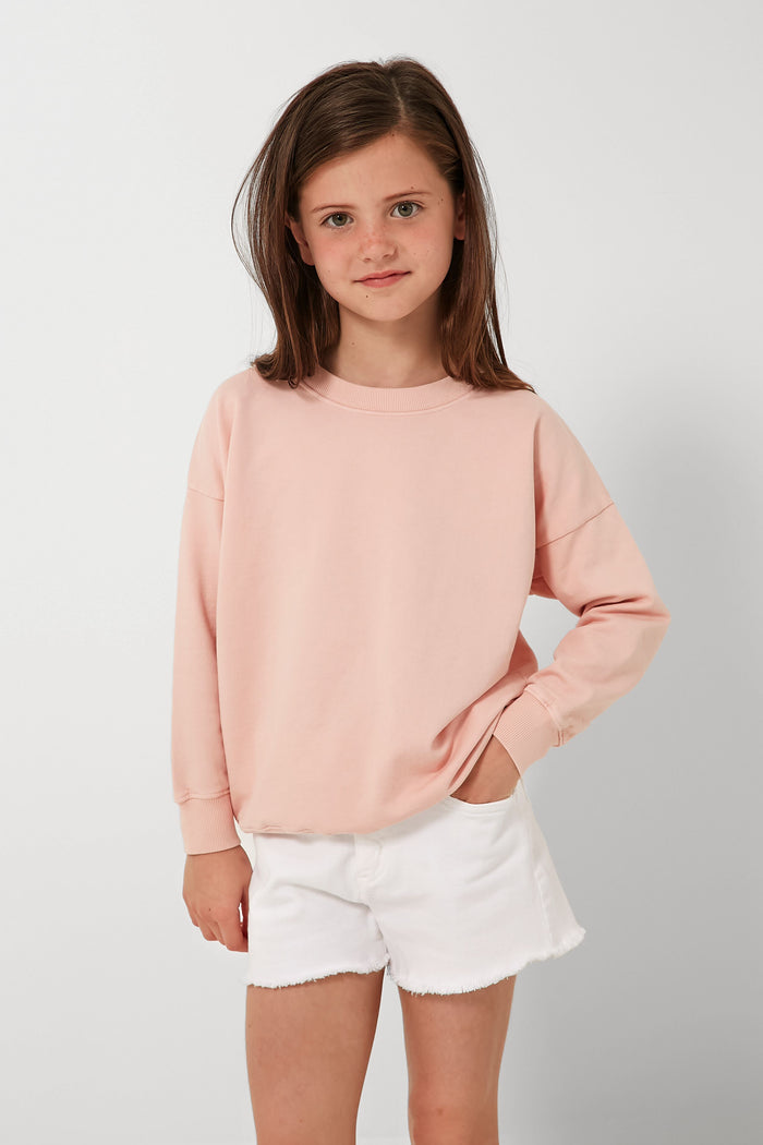 A young girl with brown hair wears a light pink organic cotton sweatshirt and white shorts from Les Gamins, with one hand in her pocket. The sustainable kids clothing is ethically made in LA.