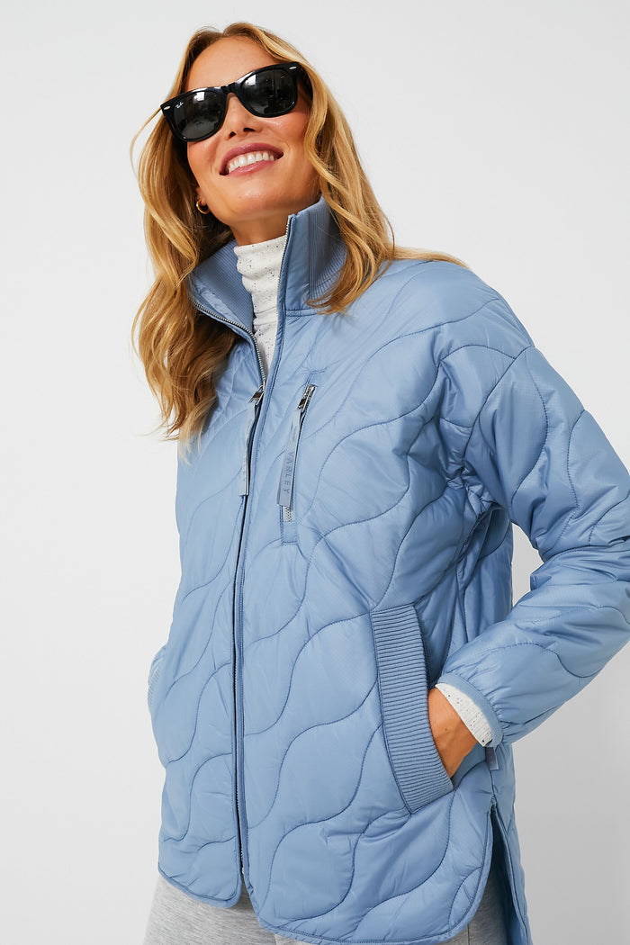 A woman smiles with her hands in her pockets, wearing a light blue Varley quilted jacket, a white turtleneck, and black sunglasses—a stylish women’s performance outerwear look by Varley.