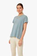 A person stands wearing a light blue THE GREAT. Boxy Crew Tee made from 100% cotton, cream-colored pants, gold hoop earrings, and a necklace.