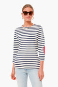 A woman in Saint James sunglasses, gold jewelry, a sailor-striped nautical shirt with red elbow patches, and light blue jeans stands confidently.