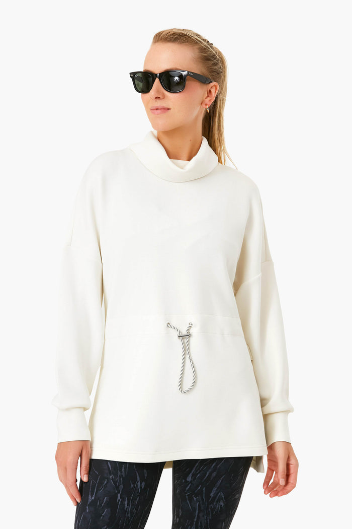 A woman models chic performance-fashion style in Varley sunglasses, a white long-sleeve cinched-waist pullover with drawstring, and dark patterned pants.