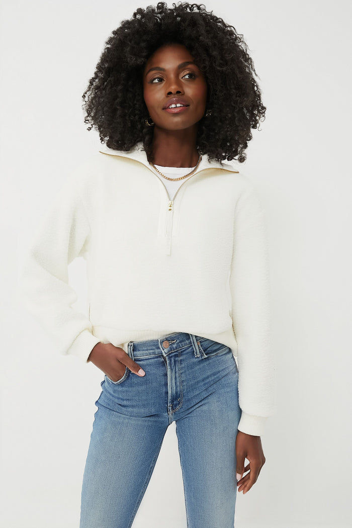 A woman wears a white Varley Egret Roselle Half Zip Fleece pullover and light blue jeans, one hand in her pocket and the other by her side, embodying effortless performance-fashion.