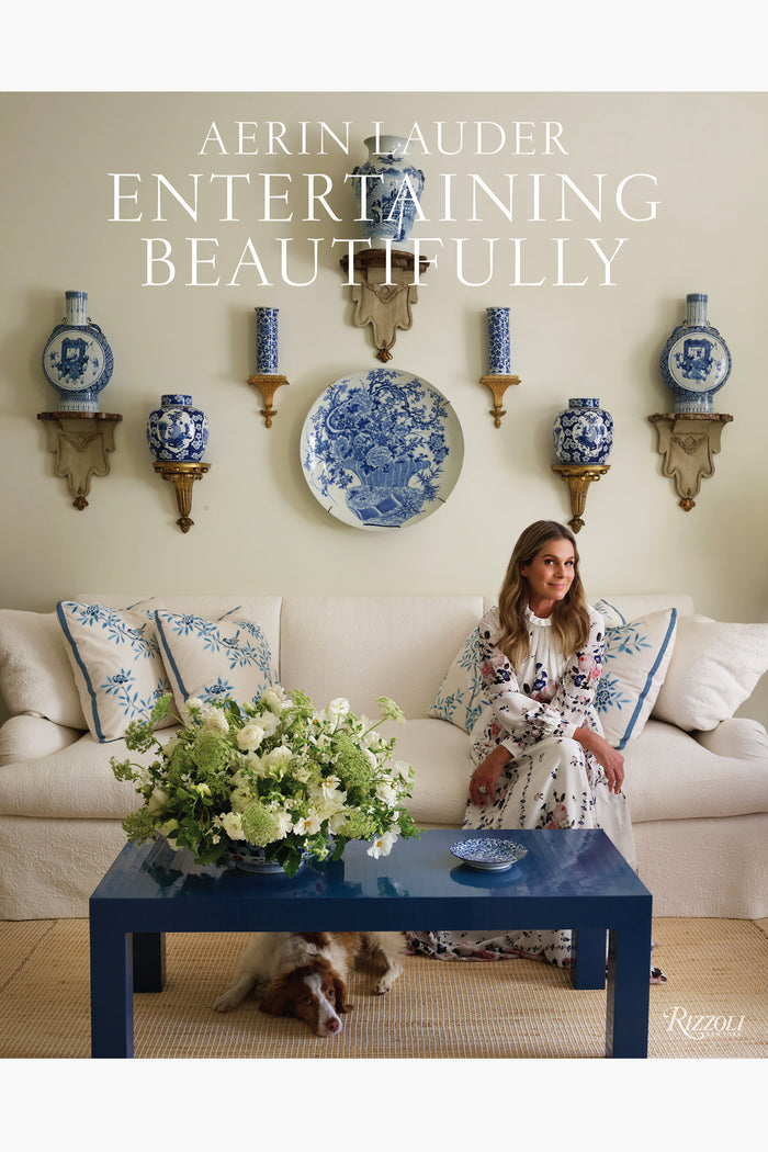 A woman in a floral dress relaxes on a sofa with blue and white pillows, surrounded by blue china; inspired by "Entertaining Beautifully" from Rizzoli, her dog rests beneath the coffee table.