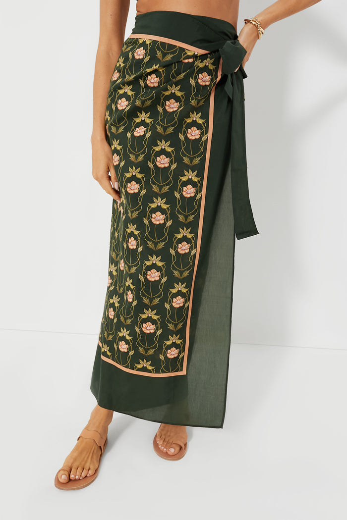 A person wears an AGUA BY AGUA BENDITA dark green floral wrap skirt with a waist tie, paired with brown sandals.