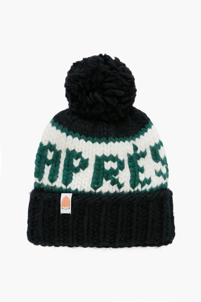 A black hand-knit merino wool beanie by Lele Sadoughi with a pom-pom, white and green stripes, and "APRES" in green across the front.