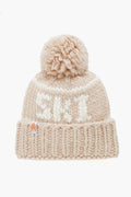 A beige Lele Sadoughi knit beanie made from Peruvian Merino wool, featuring a large pom-pom, white front lettering, and a small patch on the ribbed cuff.