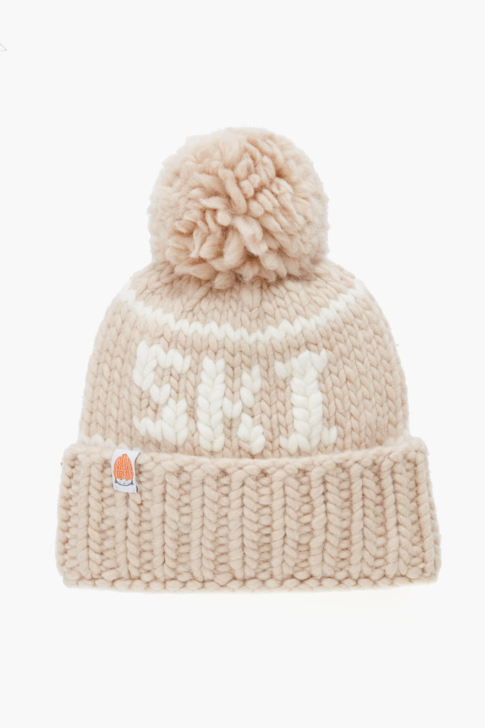 A beige Lele Sadoughi knit beanie made from Peruvian Merino wool, featuring a large pom-pom, white front lettering, and a small patch on the ribbed cuff.