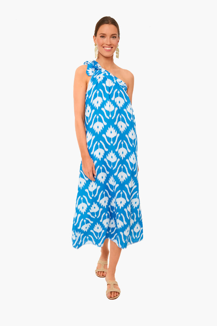 A woman wears the Nimo With Love Ikat Blue Water Lilly Dress, an elegant boho-chic one-shoulder blue-and-white midi, styled with beige sandals and drop earrings.