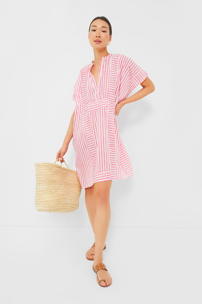 A woman wears a pink and white striped short dress from Love Tomas by Emerson Fry, tan sandals, and carries a woven straw bag.