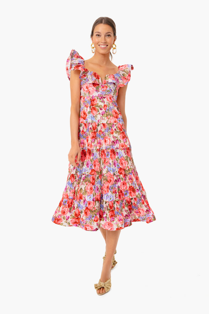 A woman stands smiling in a Beulah colorful floral dress with ruffled sleeves and a tiered A-line skirt, paired with gold hoop earrings and gold open-toe heels.