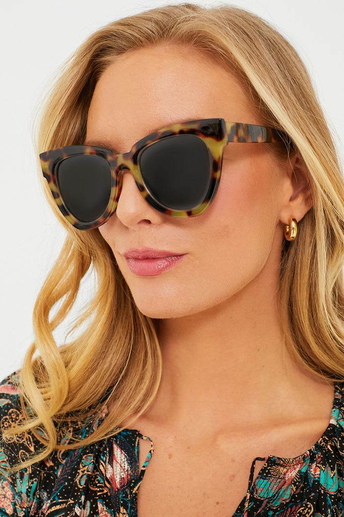 Woman wearing Exclusive Tortoise Not Standard Sunglasses, featuring oversized tortoiseshell frames and dark lenses, paired with a fun, patterned top and gold hoop earring.