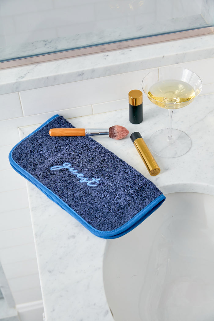 A Weezie for Tuckernuck luxury bath towel labeled "guest" sits on a marble bathroom counter with makeup items and a glass of white wine.