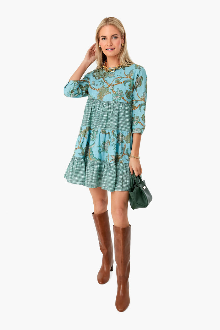 A woman wears the Ro’s Garden Turquoise Camilla Rene Dress—a blue patterned, tiered block-printed dress—with brown knee-high boots and a small green handbag.