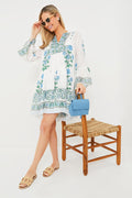 Wearing a Juliet Dunn white and blue floral block print dress, a woman stands by a woven chair with a blue handbag, sandals, and sunglasses—an ideal luxury resort look.