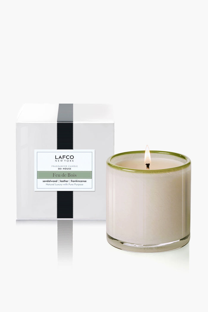 A white glass candle from LAFCO New York is lit beside its box, labeled Feu de Bois, and features home fragrances of sandalwood, leather, and frankincense.