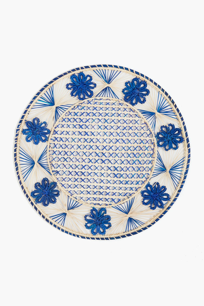 A Coro Cora round ceramic plate with a blue and white geometric pattern and blue floral rim, elegantly paired with handmade iraca palm placemats crafted by women artisans.