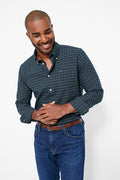 A man smiles and looks down, dressed in a dark green plaid Barbour shirt, blue jeans, and a brown belt.