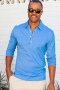 A man wears Criquet sunglasses, a blue long-sleeve men's golf shirt, and beige pants, standing with one hand in his pocket and smiling.