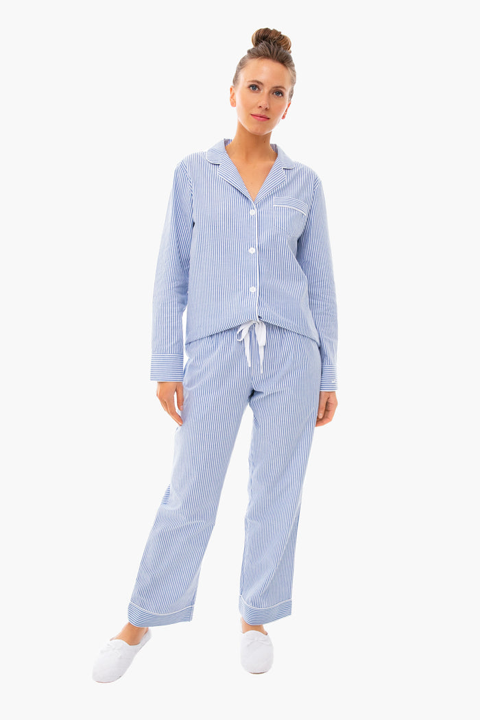 A woman wears the Petite Plume French Blue Seersucker Pajama Set with white slippers, highlighting classic striped pajamas and luxury cotton sleepwear.