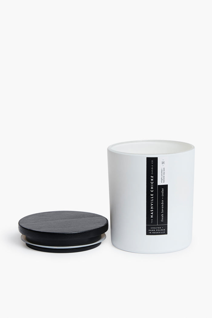 A white candle jar from The Nashville Chicks Candle Co. with a black label sits open beside its black lid, showcasing a hypoallergenic natural lavender cedar candle.
