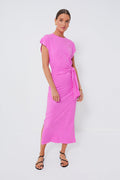 A woman wears the Apiece Apart Fuchsia Vanina Dress—an organic cotton, ankle-length dress in bright pink with short sleeves, a side tie, and black strappy sandals.