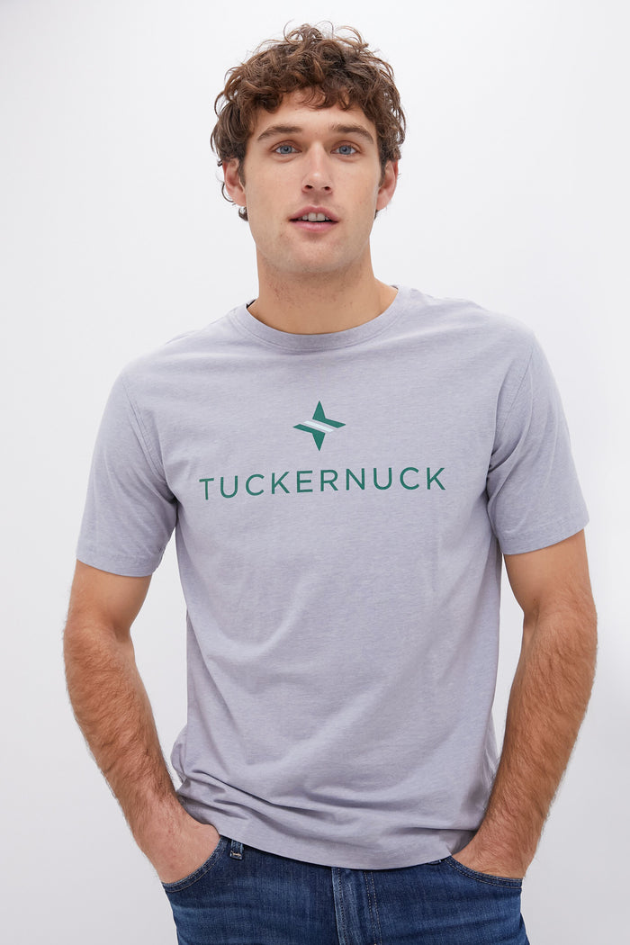 A young man with curly brown hair wears a light gray Peter Millar moisture-wicking tee with a green logo printed on the front.