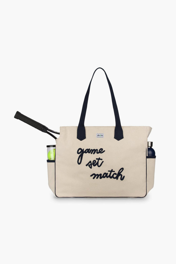 Ame & Lulu’s beige tennis tote with black handles and "game set match" front holds a racket, balls, and water bottle in side pockets—the perfect stylish bag for your court essentials.