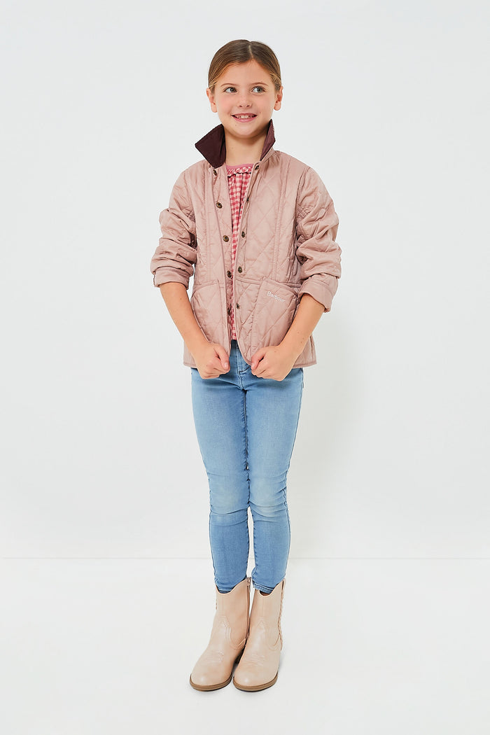 A young girl wears a light pink Barbour girls quilted jacket over a red checkered shirt, paired with blue jeans and beige boots.