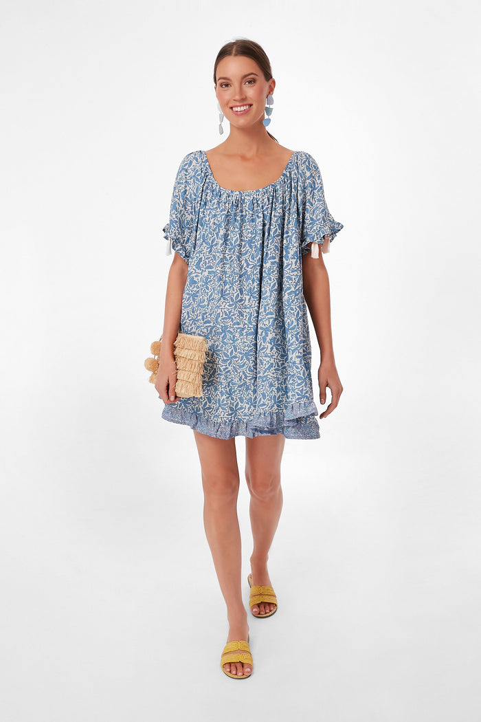 A woman in a loose blue floral batik print dress by Natalie Martin, yellow sandals, and a woven clutch highlights the charm of handmade garments.