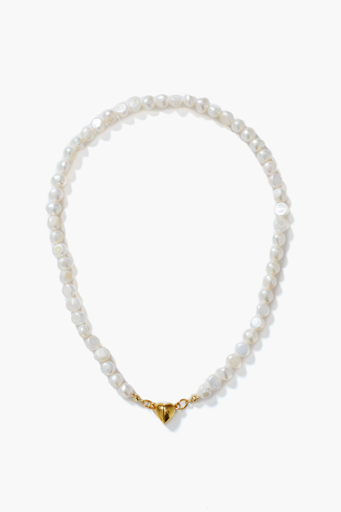A Petit Moments white pearl necklace with gold-tone hardware and a heart-shaped magnetic closure, elegantly arranged in a circle.