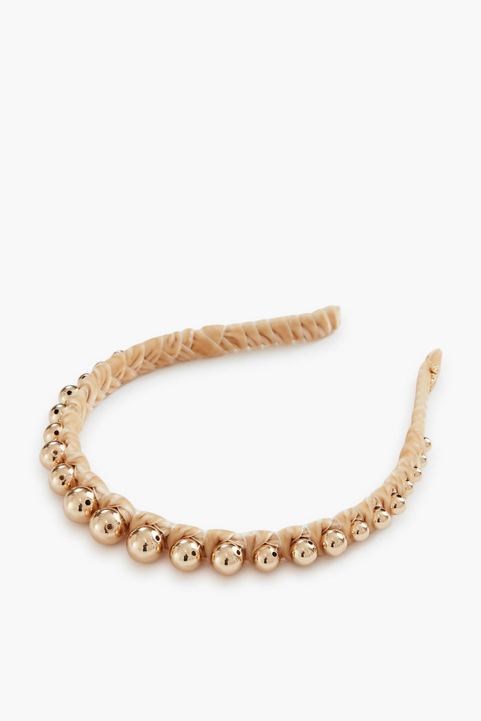 The Lele Sadoughi knotted headband is wrapped in beige fabric and features a row of gold metallic beads along the top.