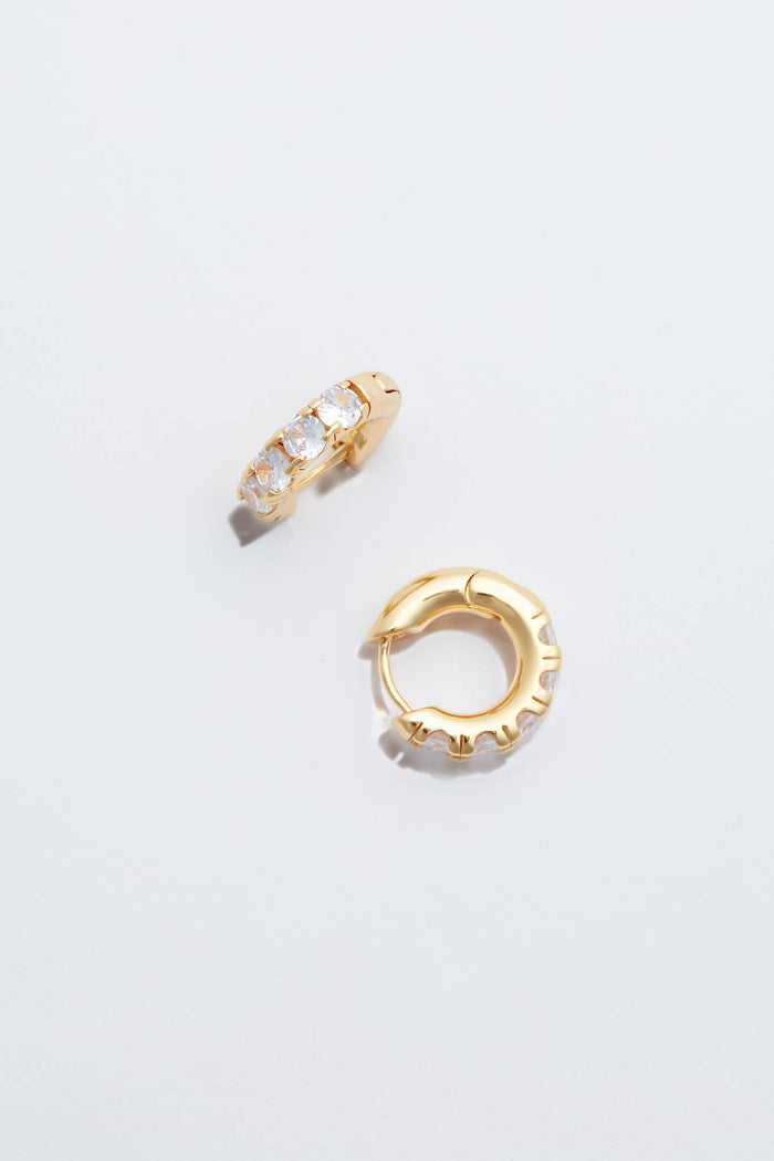 A pair of small Tuckernuck Jewelry Gold Huggie Hoop Earrings, one with clear gemstones and one plain, displayed on a white surface.