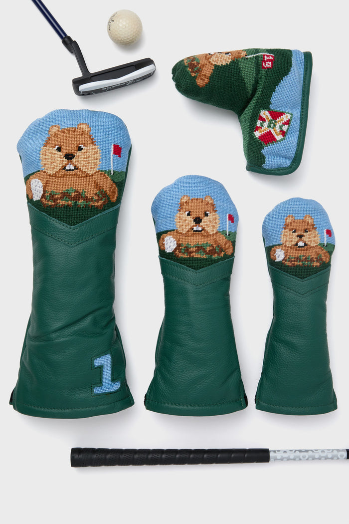 A set of Smathers and Branson golf accessories—including three green dog-embroidered headcovers, a hand-stitched hybrid cover, green putter cover, golf ball, putter, and grip—are displayed on a white surface; perfect golf gifts.