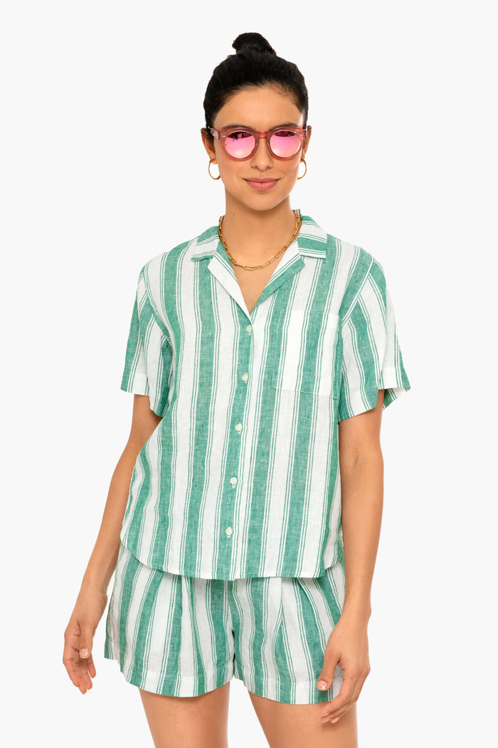 A woman exudes confidence in pink sunglasses and a matching TROVATA Birds of Paradis Green Awning Stripe Margot Shirt and shorts set—a chic, green-and-white striped short-sleeve button-up ensemble.