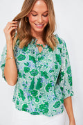A woman smiles and looks down, wearing a green and white floral bohemian shirt with tassels from Ro's Garden, styled with white pants and gold jewelry—an ideal summer look.