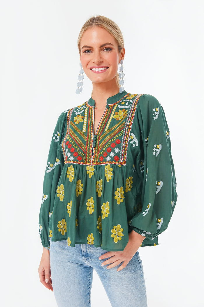 A woman wears an Oliphant green bohemian blouse with floral patterns and colorful embroidery, paired with light blue jeans and drop earrings.