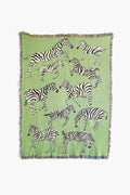 The Olivia Wendel Green Zebras Blanket is a lightweight throw made from 100% US-grown cotton, featuring playful black and white zebras in various poses on a green background.