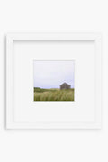 Photograph by Lauren Marttila featuring a small, weathered wooden building in a grassy field beneath an overcast sky, set in a white square frame—perfect Nantucket art or unique bookshelf decor.