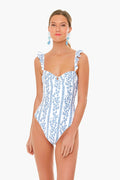 Woman wearing an AGUA BY AGUA BENDITA Colombian one-piece swimsuit with blue floral and stripe patterns, ruffled shoulder straps, and blue earrings.