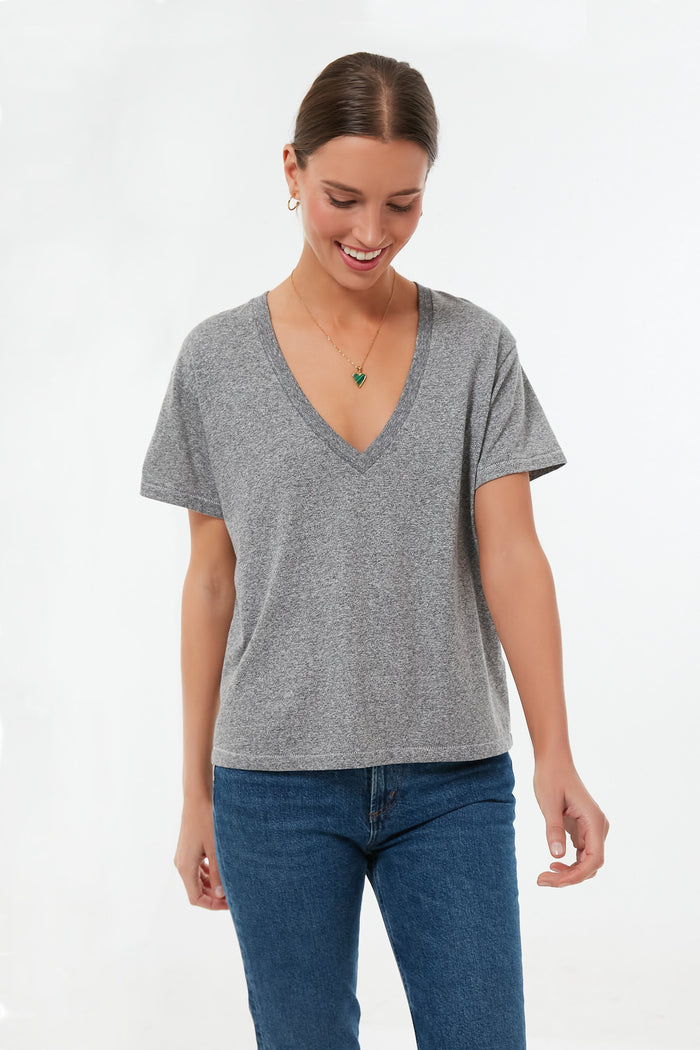 A woman in a relaxed fit THE GREAT. Heather Grey V Neck Tee and blue jeans stands, looking down with a smile.
