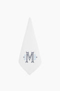 A white linen-cotton napkin by Caton's Wharf is folded into a triangle and features a decorative embroidered letter M with blue accents in the center, making it an ideal personalized gift.