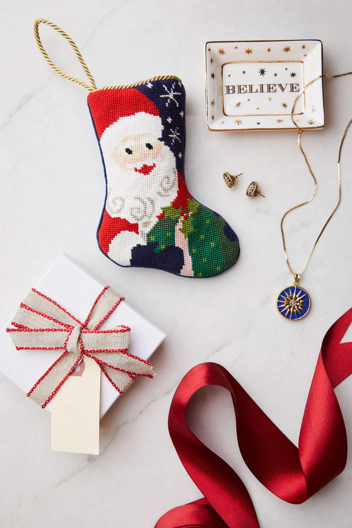 A Bauble Stockings hand-stitched Christmas stocking with Santa, a gift box, red ribbon, jewelry, and a BELIEVE dish are arranged on white—perfect fair trade gifts from Bauble Stockings this season.