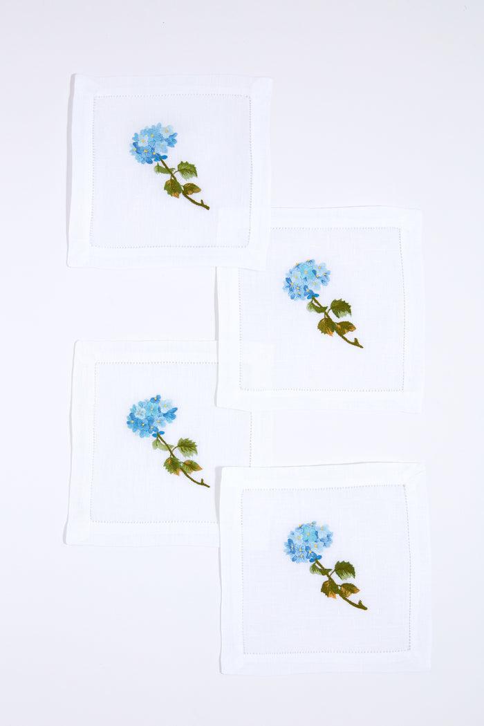 Set of four white linen napkins by Tuckernuck Home, each hand-embroidered with a blue flower and green leaves. Ideal as hydrangea-themed cocktail napkins or a thoughtful hostess gift.
