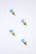 Set of four white linen napkins by Tuckernuck Home, each hand-embroidered with a blue flower and green leaves. Ideal as hydrangea-themed cocktail napkins or a thoughtful hostess gift.
