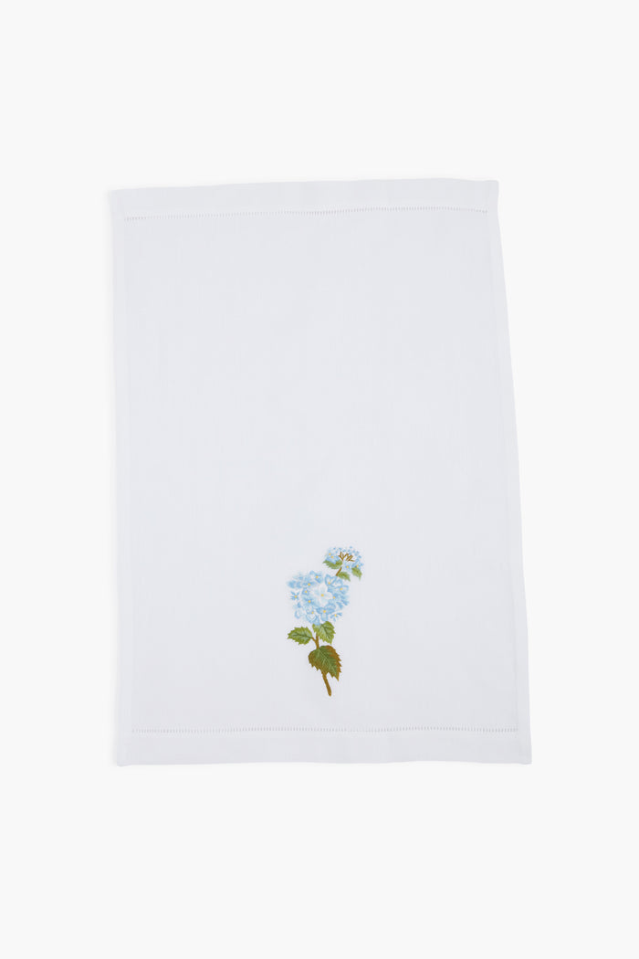 A white linen guest towel by Tuckernuck Home, featuring a hand-embroidered blue flower with green leaves in one corner.