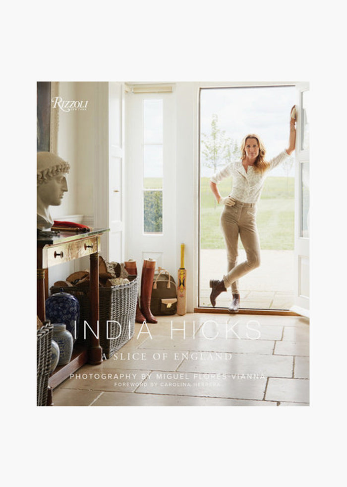 A woman stands in the doorway of a bright, elegant room with country decor. She holds "India Hicks: A Slice of England," a Rizzoli book showcasing the charm of English countryside designer homes.