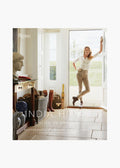A woman stands in the doorway of a bright, elegant room with country decor. She holds "India Hicks: A Slice of England," a Rizzoli book showcasing the charm of English countryside designer homes.