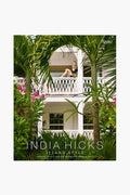 A woman relaxes on a balcony of a white two-story house with palm trees and pink flowers. The text reads, Rizzoli presents "India Hicks Island Style," showcasing relaxed island living and bohemian décor from Rizzoli.